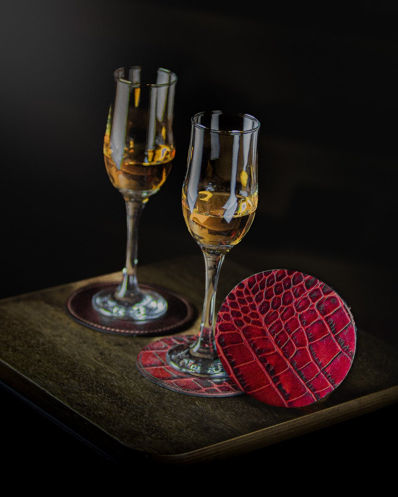 Two glasses of amber liquid on red crocodile-patterned coasters with a dark background.