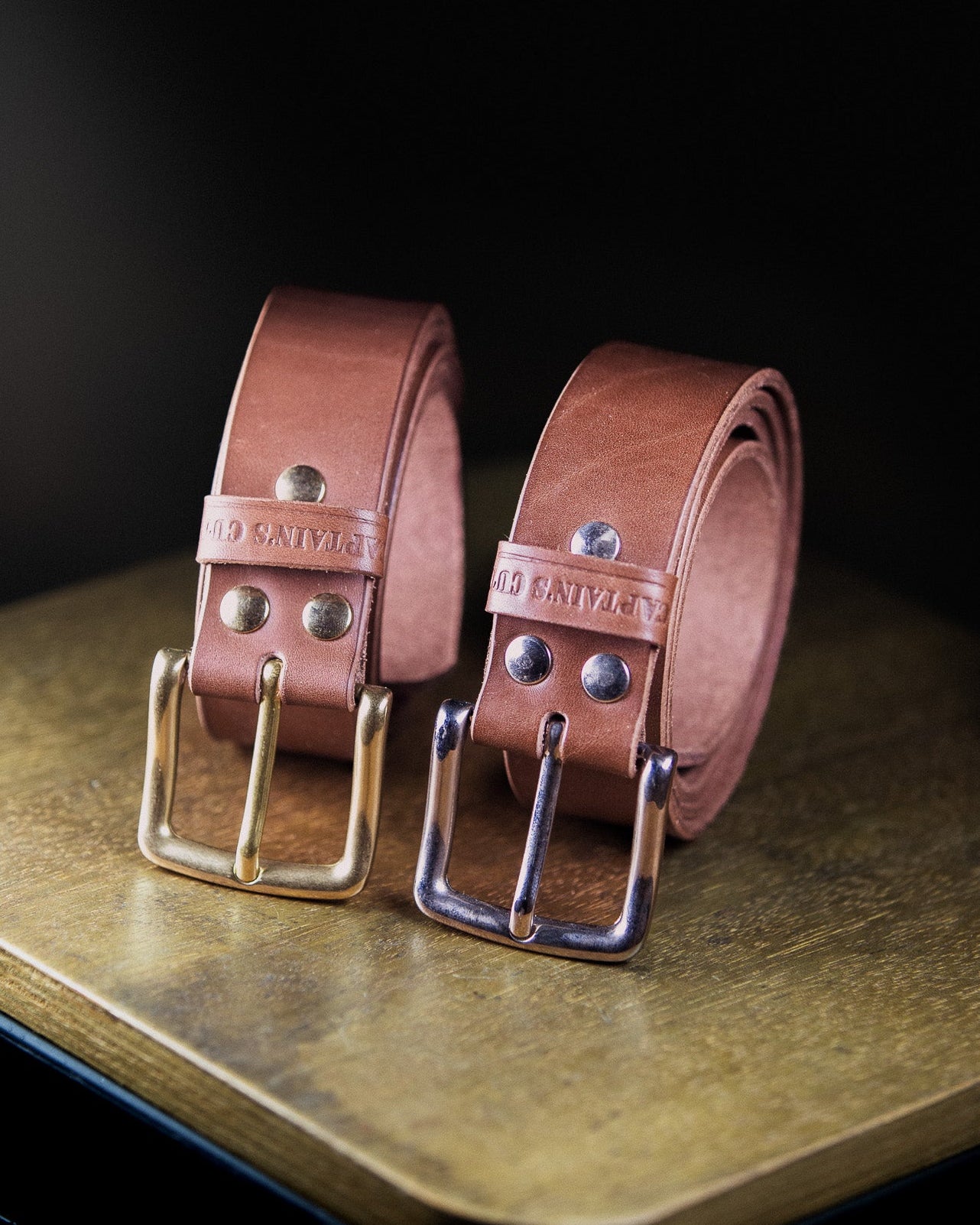 Two brown leather belts with brass buckles on a wooden surface.