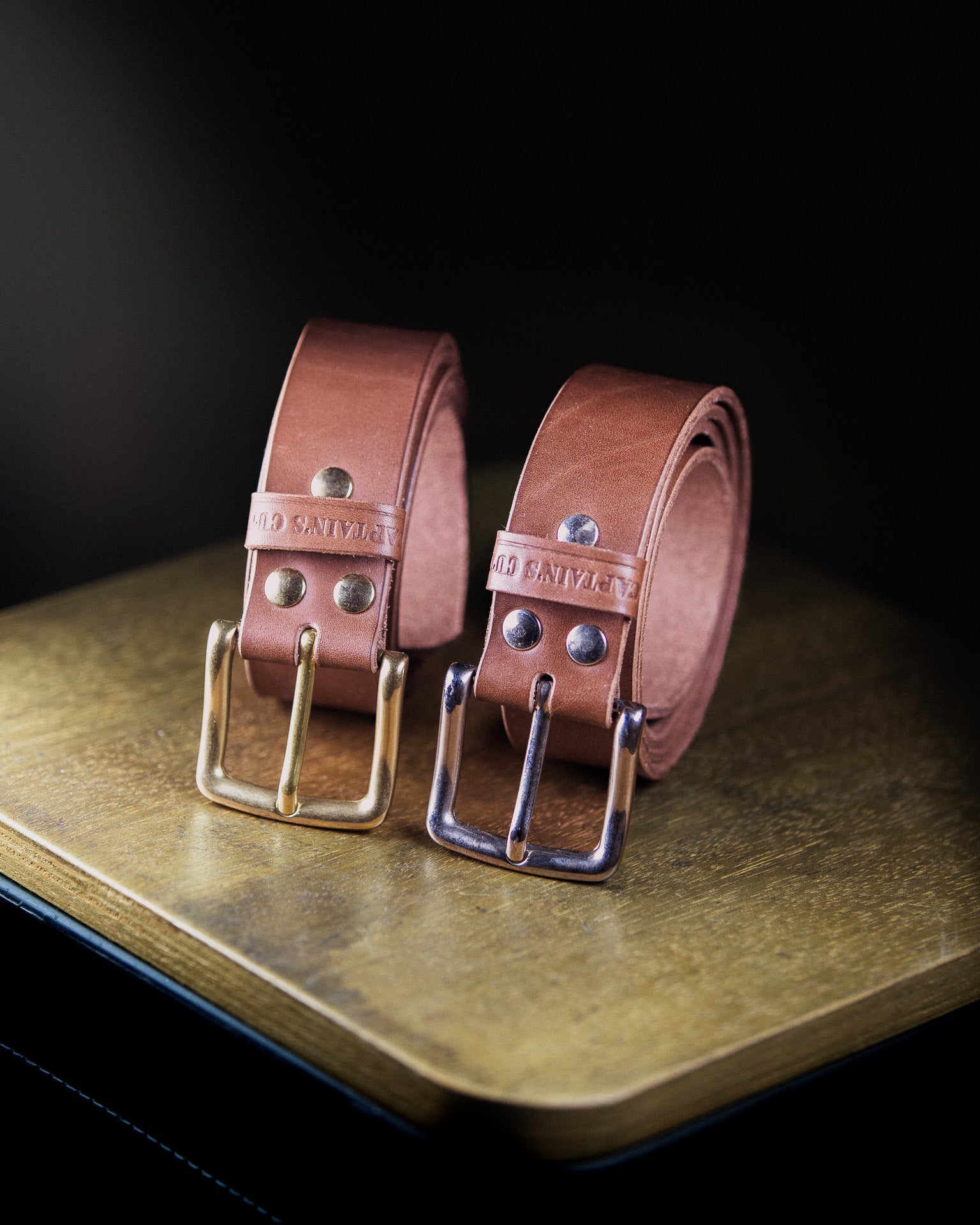 Two brown leather belts with silver  and gold buckle on a textured surface