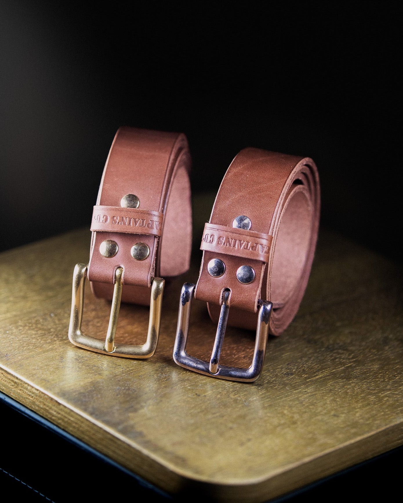 Two brown leather belts with brass buckles on a wooden surface.